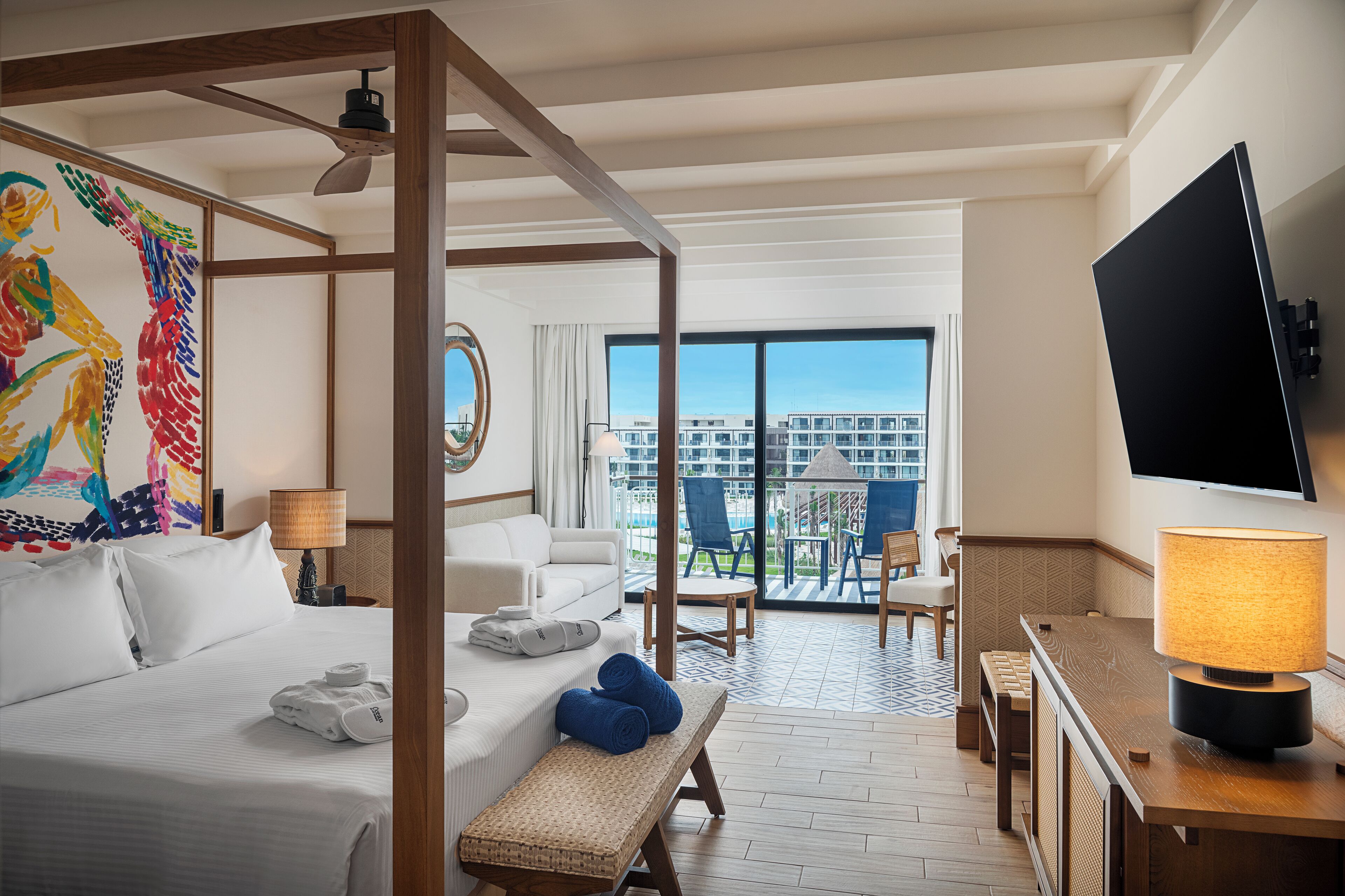 A hotel room with a bed, a bench, a desk with a lamp, a TV, a balcony with a view of buildings, and a colorful artwork on the wall.
