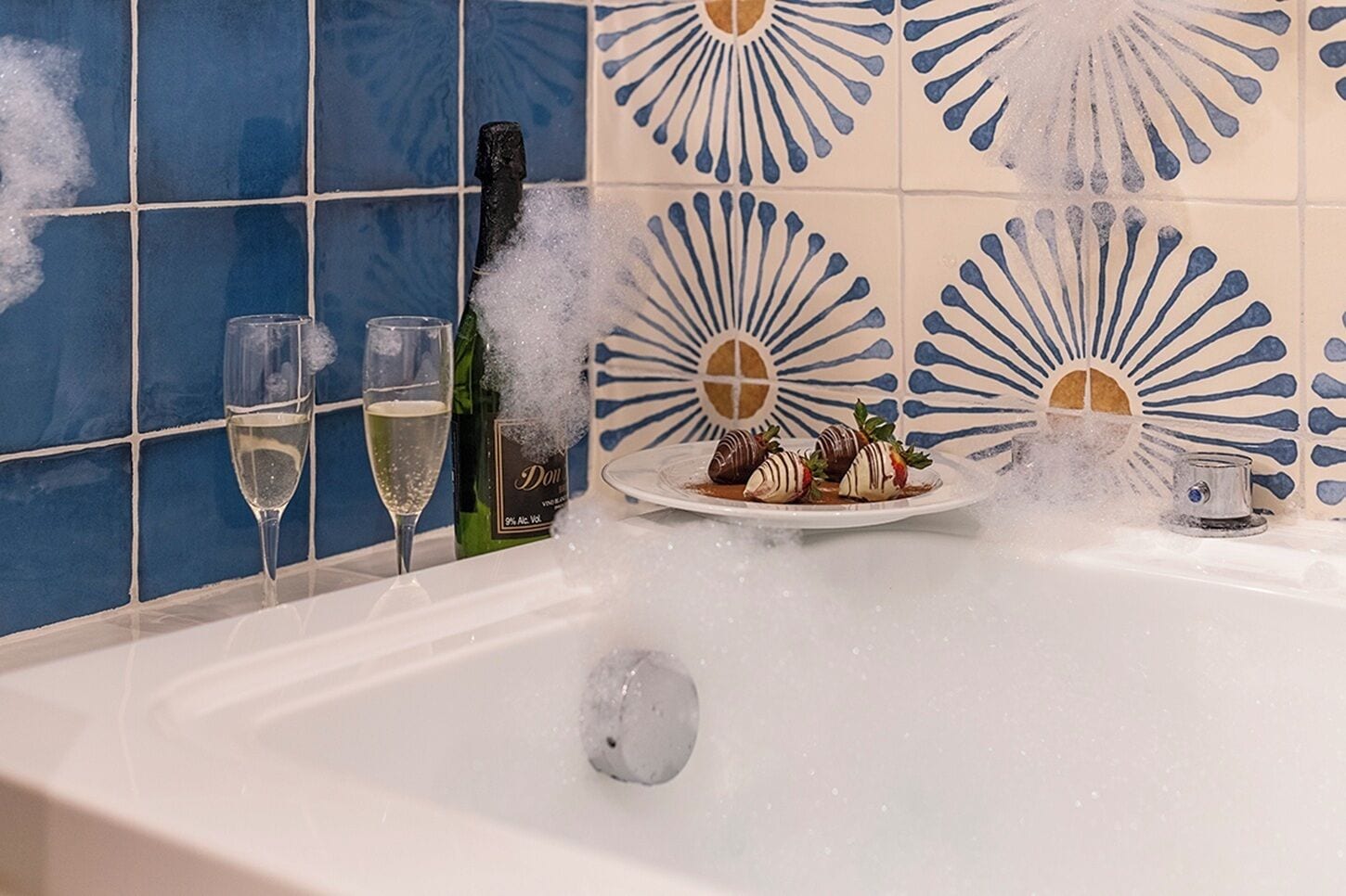 A bathtub with a champagne bottle, glasses, and chocolate-covered strawberries.