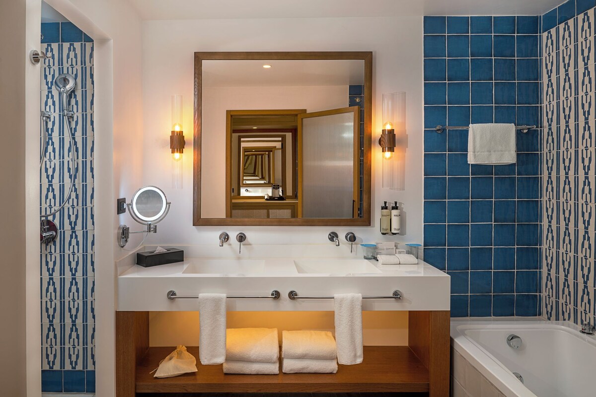 A bathroom with a large mirror, two sinks, a bathtub, and blue-tiled walls.