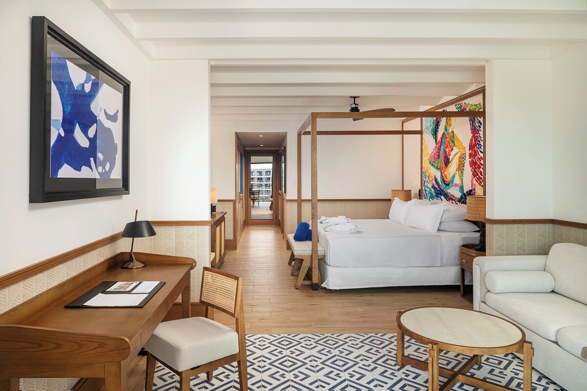 A hotel room with a bed, a desk, a chair, a small table, and a colorful artwork on the wall.