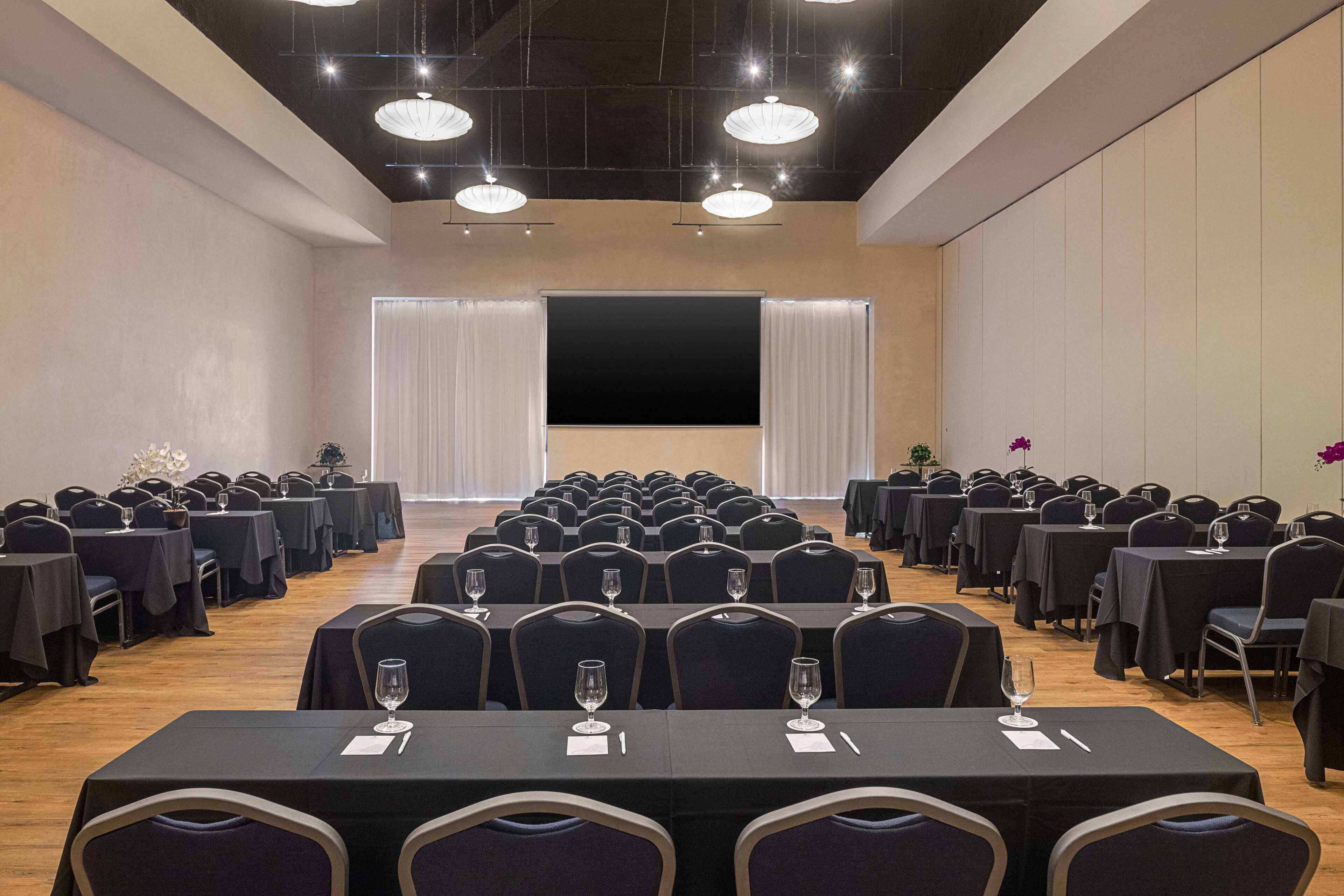 A conference room with rows of tables and chairs, a stage with curtains, and ceiling lights.