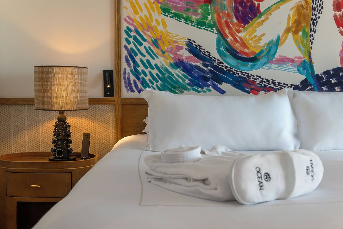 Una habitación de hotel con una cama bien hecha, una lámpara de noche, un cuadro abstracto de colores y un cabecero de madera.