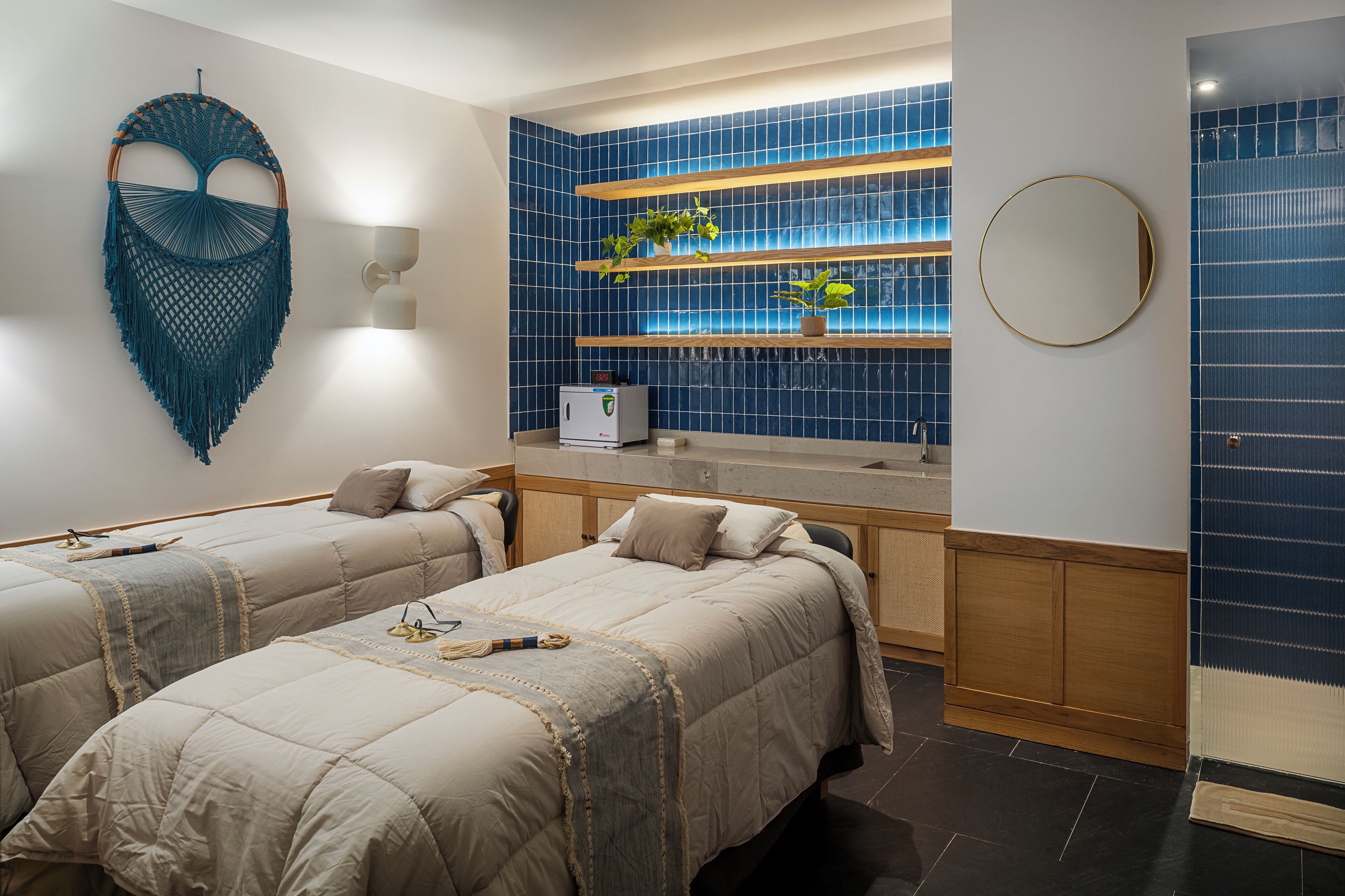 A hotel room with two beds, a wall-mounted macrame piece, a shelf with plants, and a round mirror.