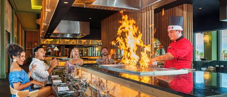 A chef performing a flambé at a restaurant counter with diners seated at a table.