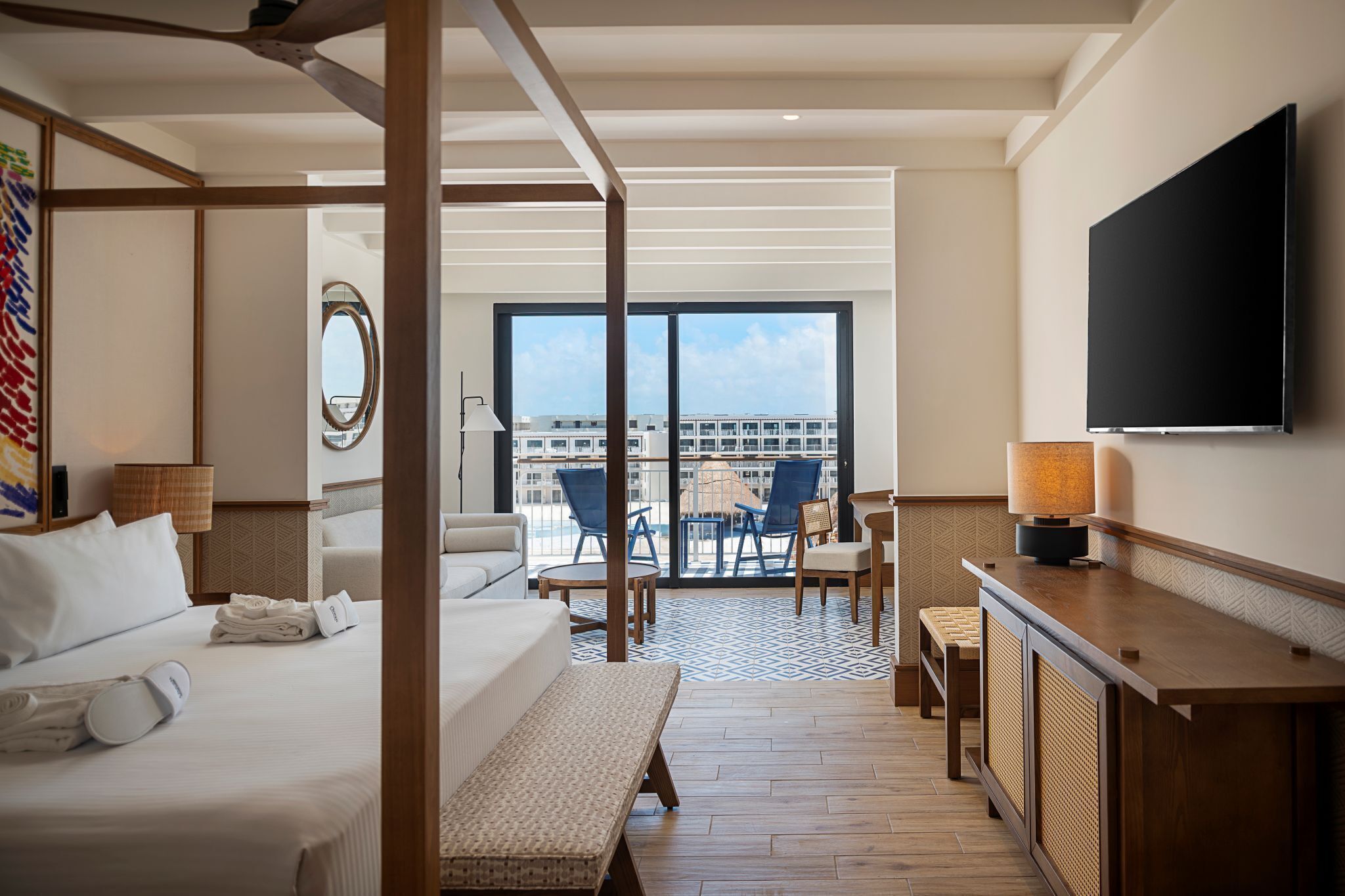 A hotel room with a bed, a desk, a TV, a balcony with chairs, and a view of the city.