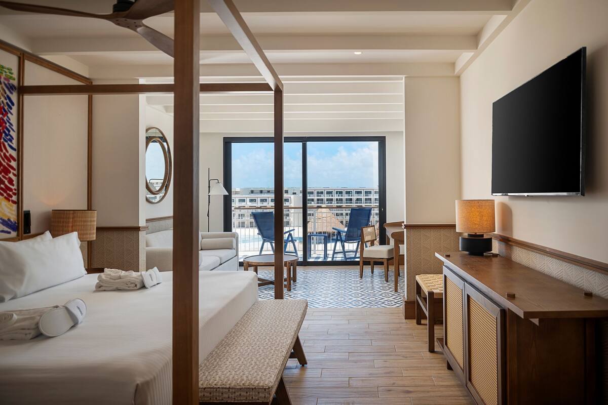 Habitación de hotel con cama, escritorio, televisión, balcón con sillas y vista a la ciudad.