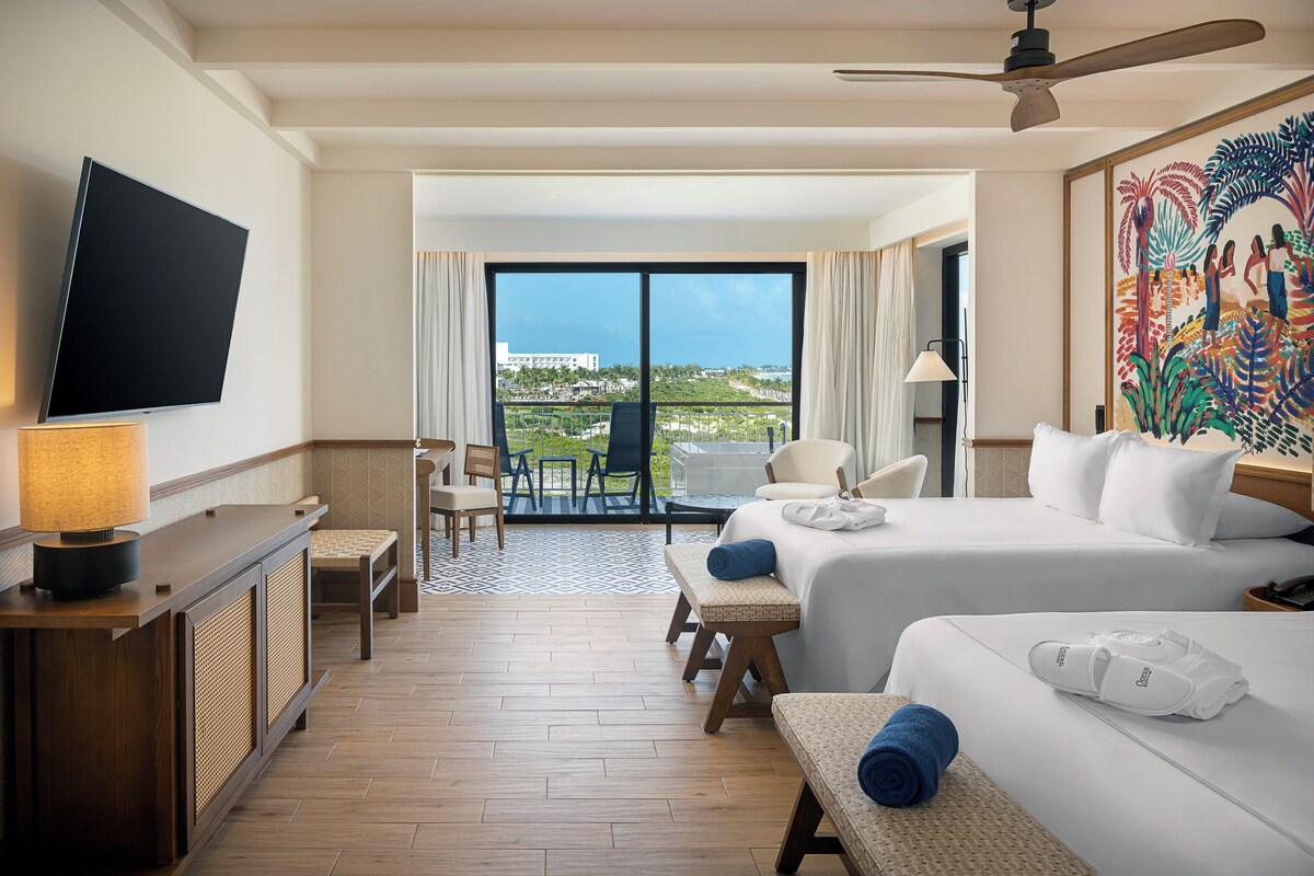 A hotel room with a TV, a desk, a chair, a bed, a balcony with a view, and a painting.