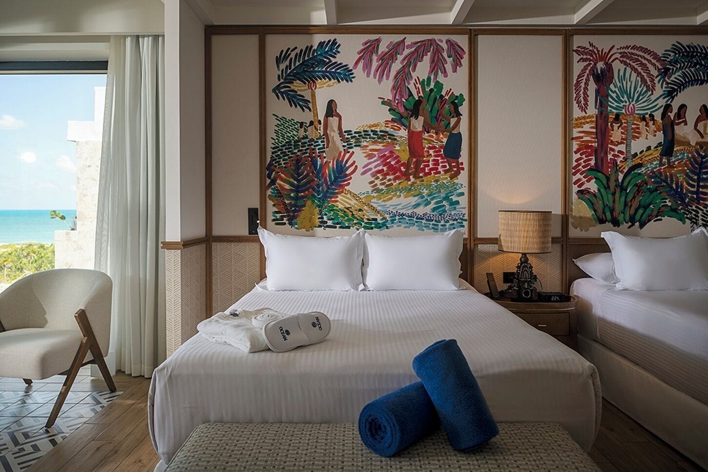 A hotel room with a bed, two pillows, a pair of slippers, a lamp, and a painting on the wall.