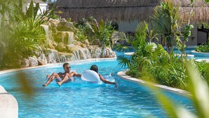 Una piscina con cascada, rodeada de plantas tropicales y una estructura con techo de paja.