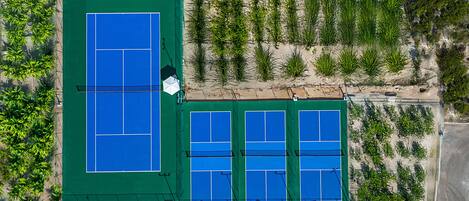 Vista aérea de un complejo de canchas de tenis con varias pistas, rodeado de vegetación y una carretera.