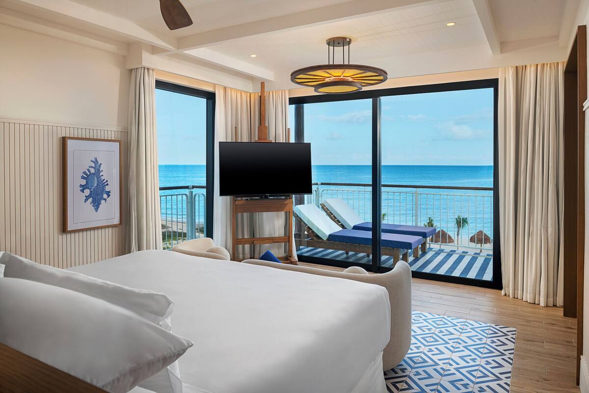 A hotel room with a bed, a TV, a balcony with lounge chairs, and a view of the ocean.