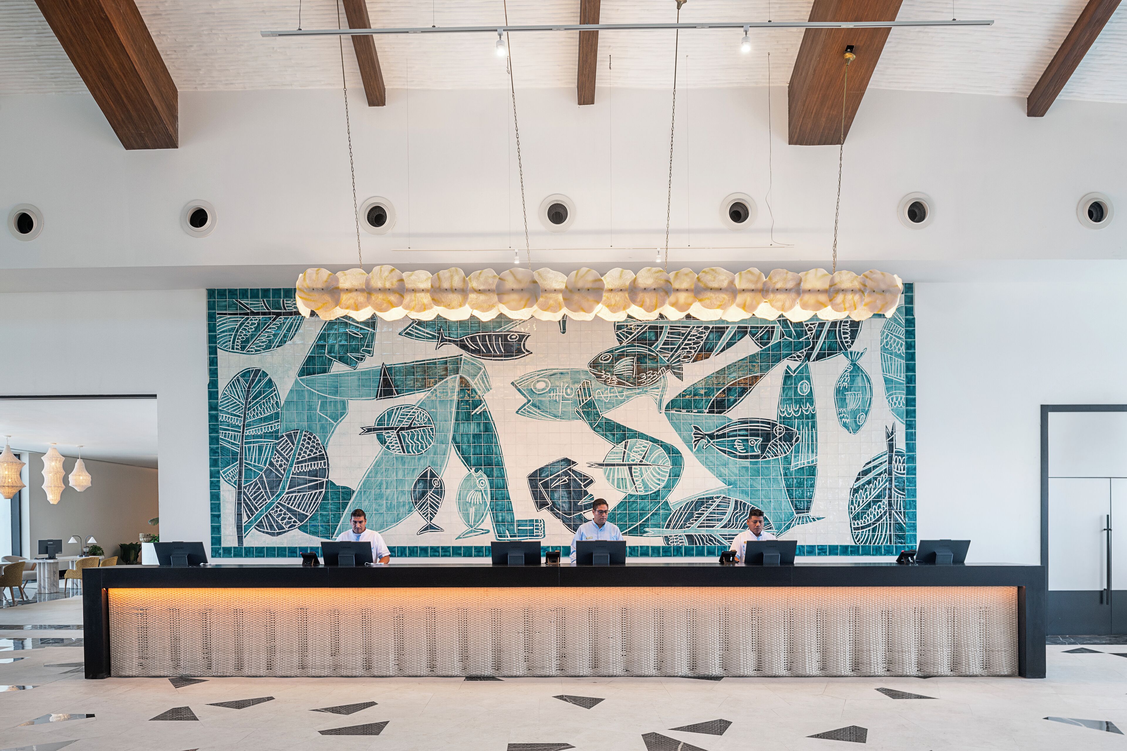 A reception desk with a mosaic wall art, a ceiling with wooden beams, and a modern chandelier.