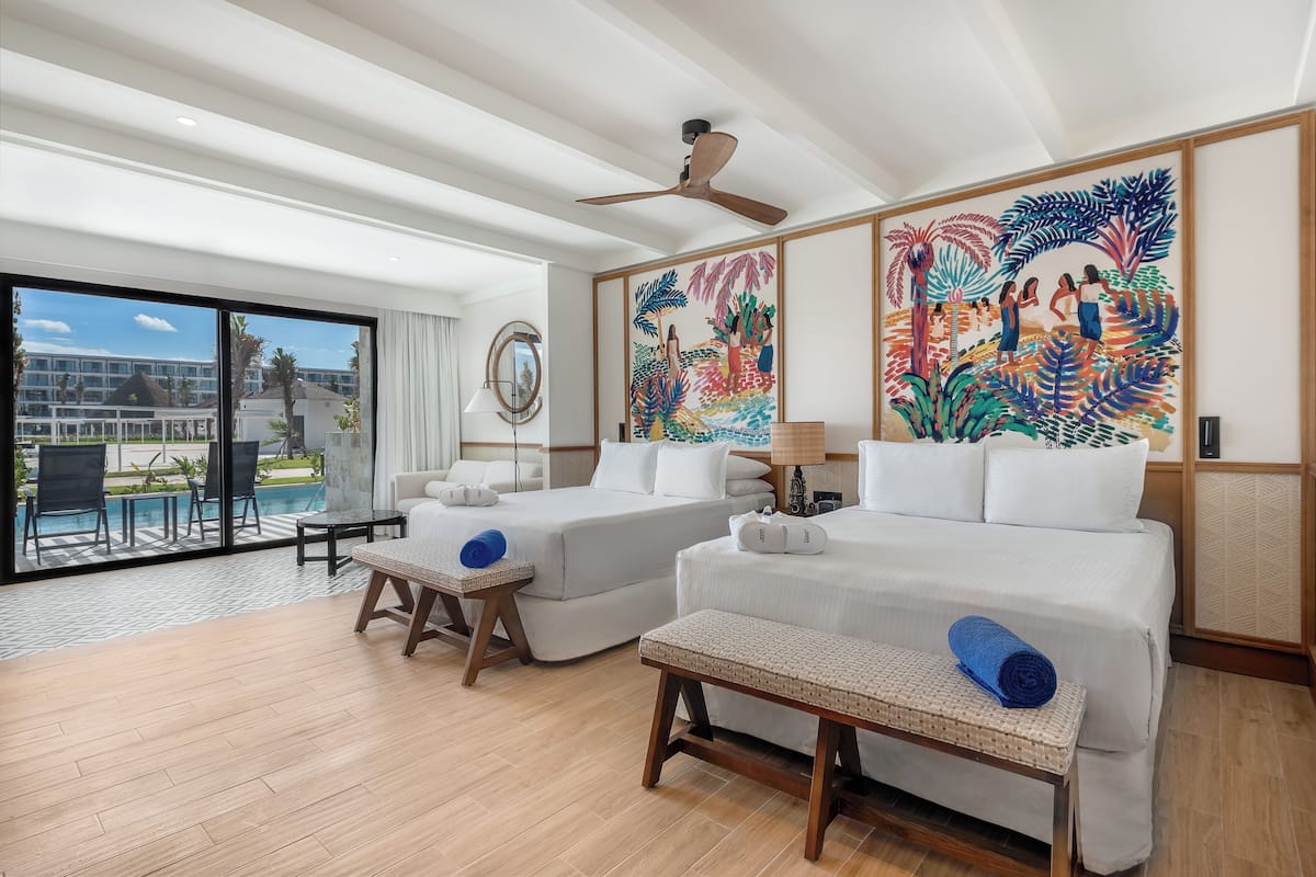 Habitación de hotel con dos camas, un ventilador de techo, un ventanal con vistas a un edificio y palmeras, y un cuadro en la pared.