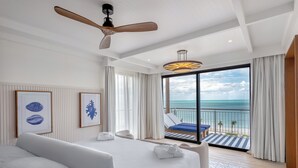 Habitación de hotel con una cama grande, ventilador de techo, balcón con vistas al océano y obras de arte enmarcadas en la pared.