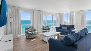 A modern living room with a large window overlooking the ocean, a blue sofa, a patterned armchair, and a white coffee table. - 🌅 Gulf Front + Seasonal Comp Beach Setup | Shoreline 1081 - Stunning Views (Destin)