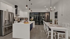 Una cocina moderna con una isla central, armarios blancos, electrodomésticos de acero inoxidable e iluminación colgante, además de un comedor con sillas blancas.