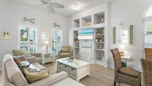 Een woonkamer met een beige bank, een glazen salontafel, rieten stoelen en een televisie bevestigd aan een witte kast.