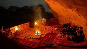 Un grupo de personas sentadas alrededor de una fogata en un espacio parecido a una cueva, con una gran roca sobre ellas.