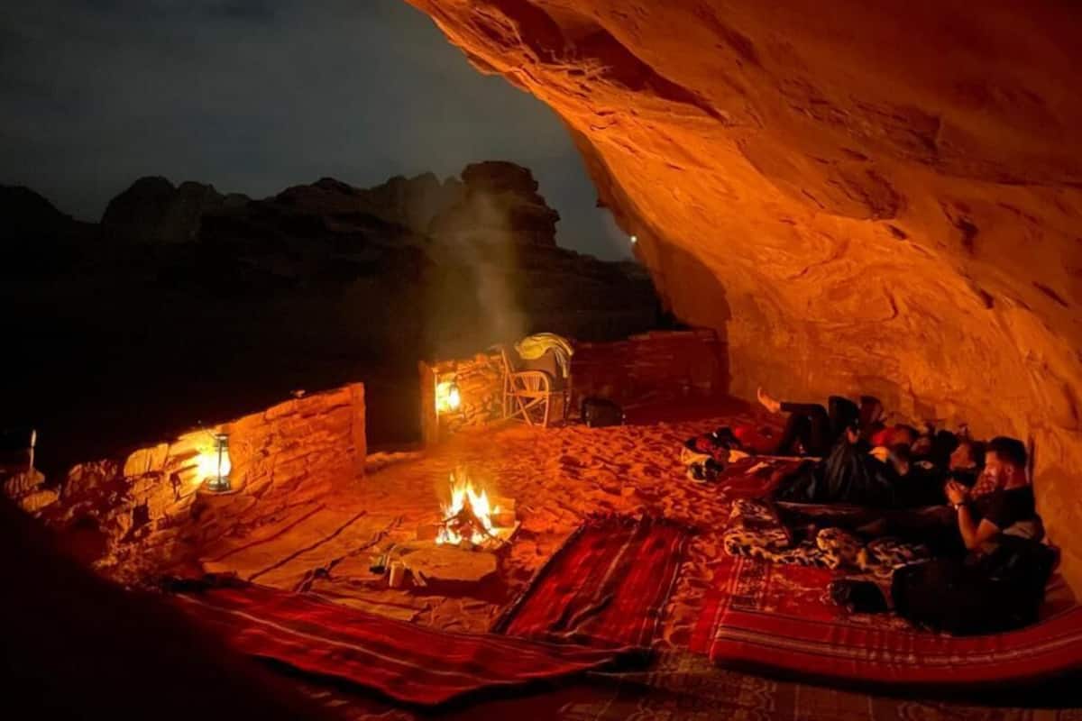 A group of people sitting around a fire in a cave-like setting with a large rock overhead.