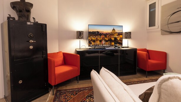 A hotel room with a large TV, two red armchairs, a black safe, and a view of a cityscape.