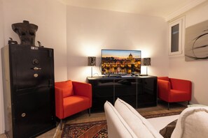 Habitación de hotel con un televisor grande, dos sillones rojos, una caja fuerte negra y vista a un paisaje urbano.