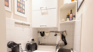 A compact kitchen with a dishwasher, a coffee maker, and a small sink. There are framed pictures on the wall and a shelf with kitchen items.