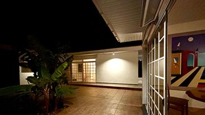 Un patio con suelo de baldosas, un ventanal y una puerta. Se ven plantas y un banco.