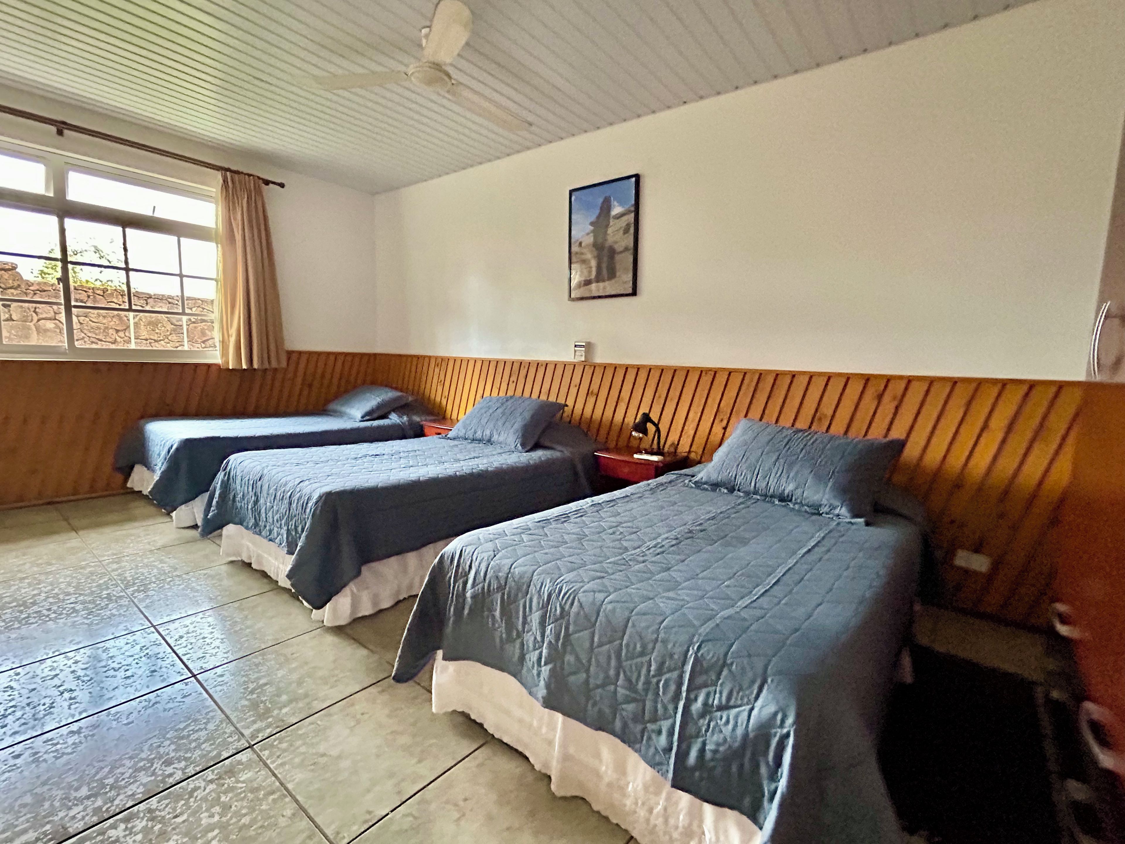 A room with three beds, a ceiling fan, a window with curtains, and a wall-mounted picture.