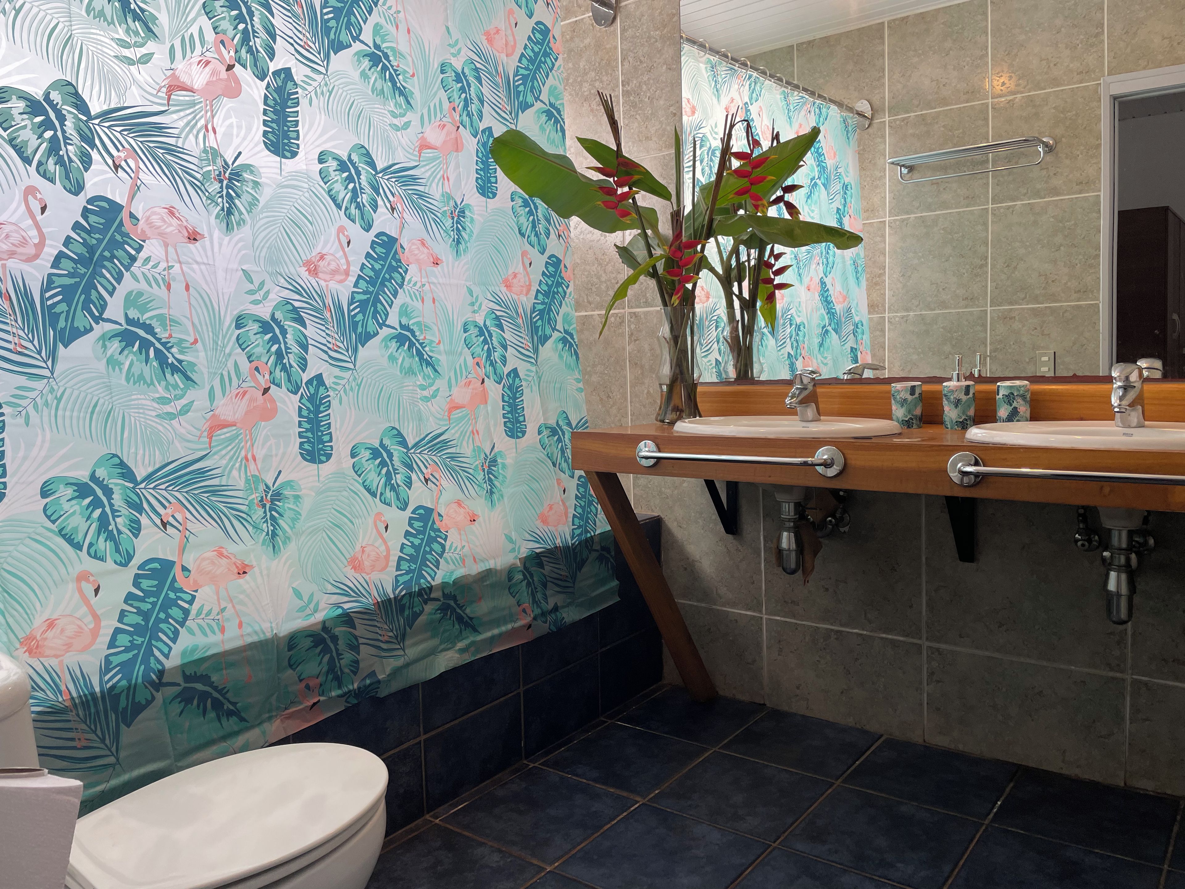 Un baño con inodoro, lavamanos con espejo y cortina de ducha con estampado de hojas tropicales y flamencos.