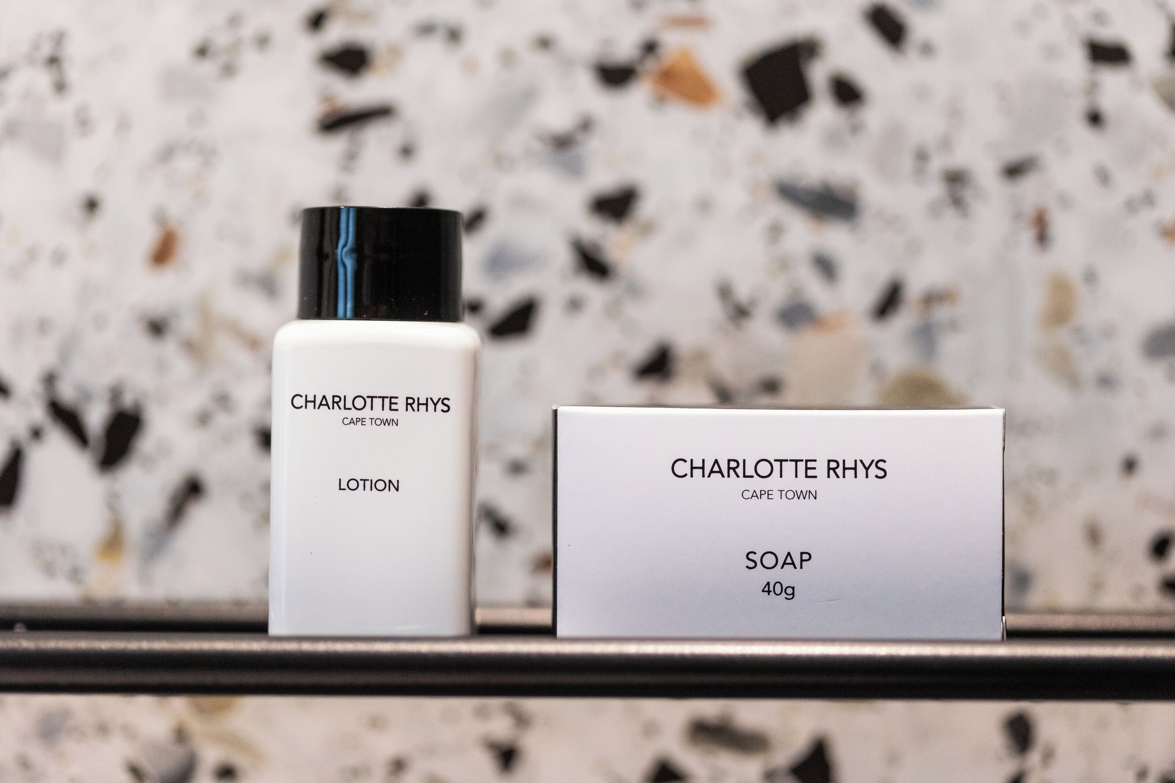 Two Charlotte Rhys products, a lotion and a soap, on a shelf against a speckled background.