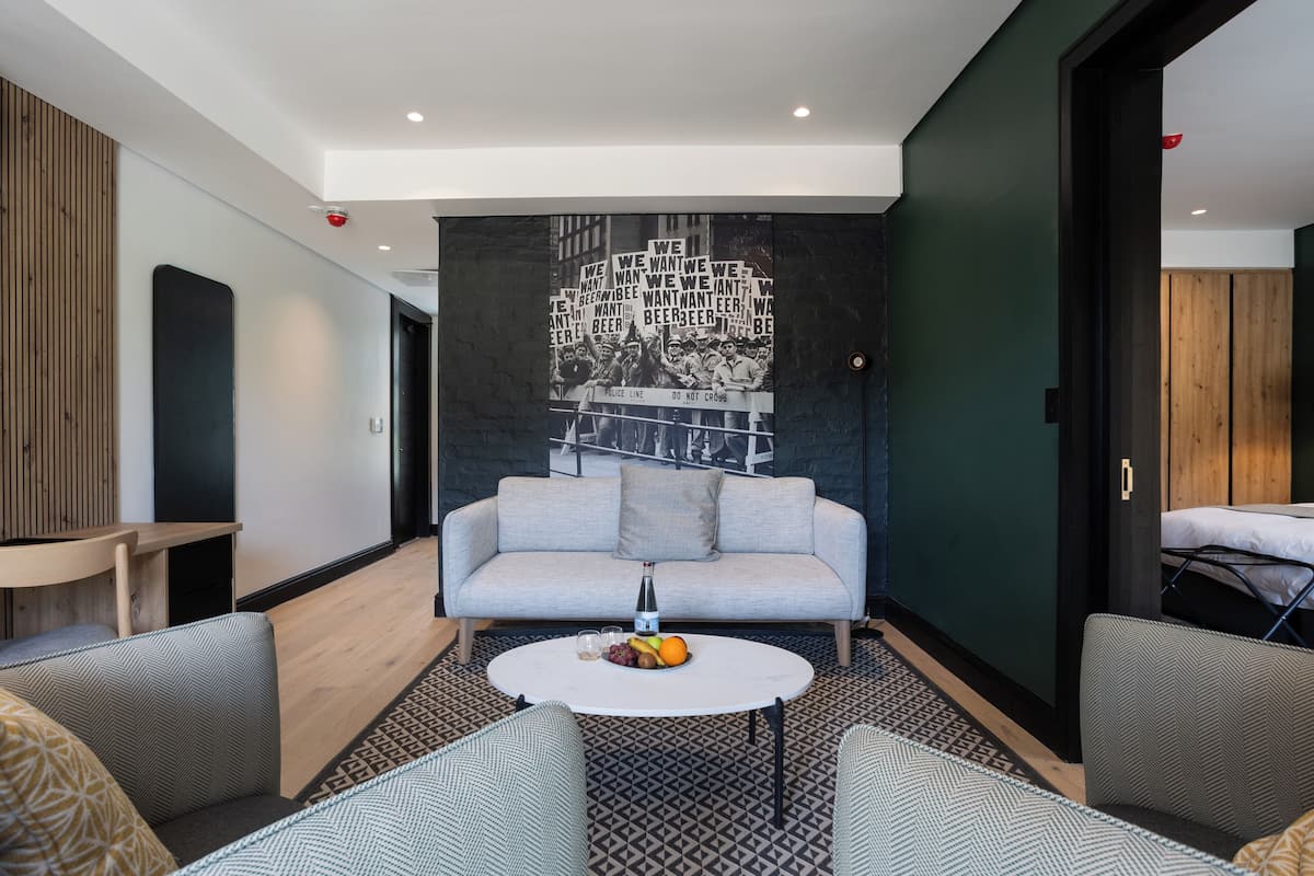 Ein modernes Hotelzimmer mit grauem Sofa, rundem Couchtisch mit Früchten und Wandgemälde.