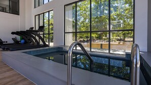 A modern indoor swimming pool with clear glass walls, exercise equipment, and a view of a park.
