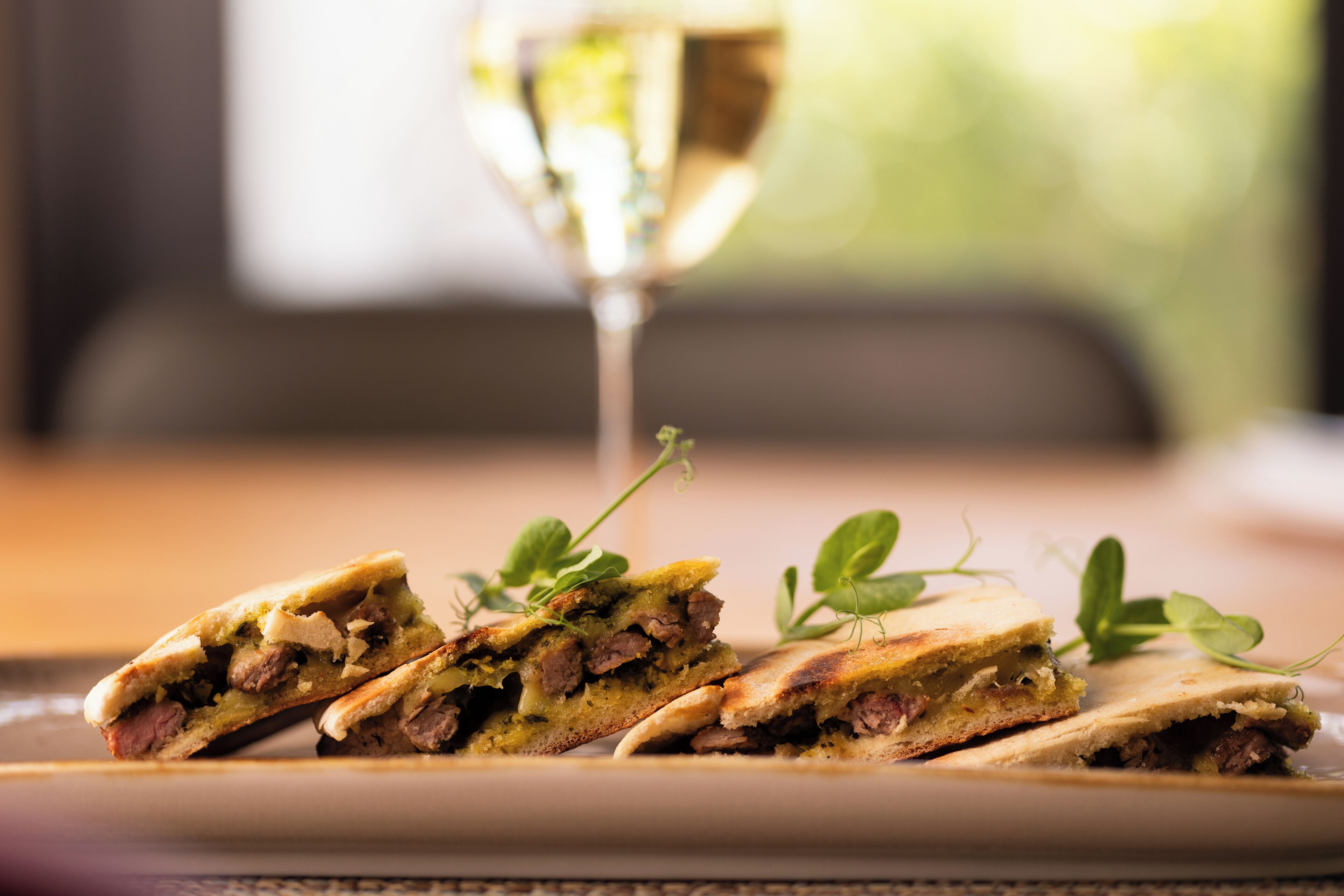 A plate with a sandwich cut in half, garnished with a sprig of herb, and a glass of white wine in the background.
