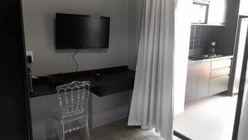 Quarto com televisão de tela plana presa na parede, uma escrivaninha com cadeira e área de cozinha visível por uma porta aberta.