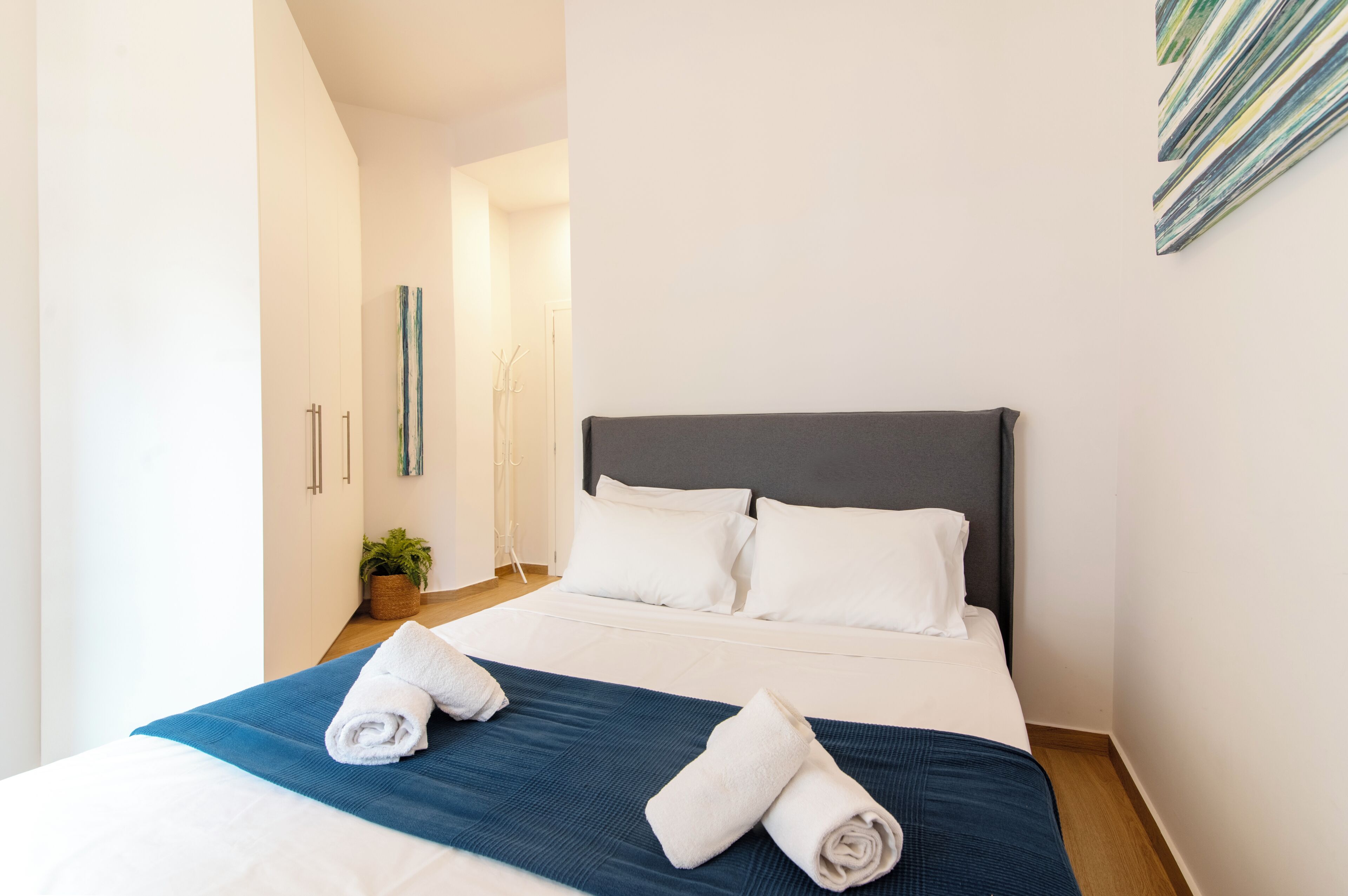 A neatly made bed with white and blue bedding, a dark headboard, and two rolled towels on the bed.