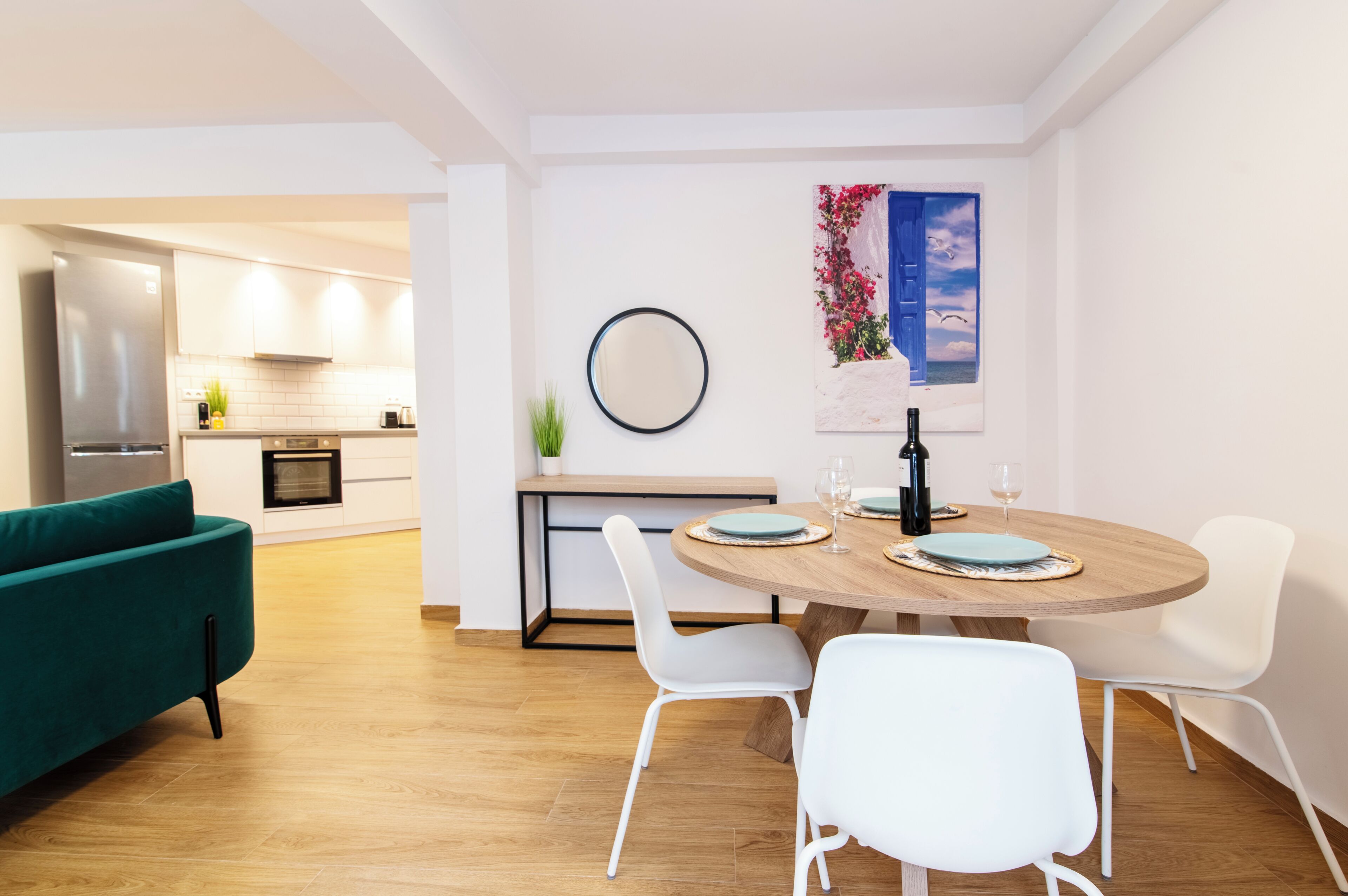 A modern kitchen with a built-in oven, a dining area with a round wooden table and white chairs, and a teal sofa.