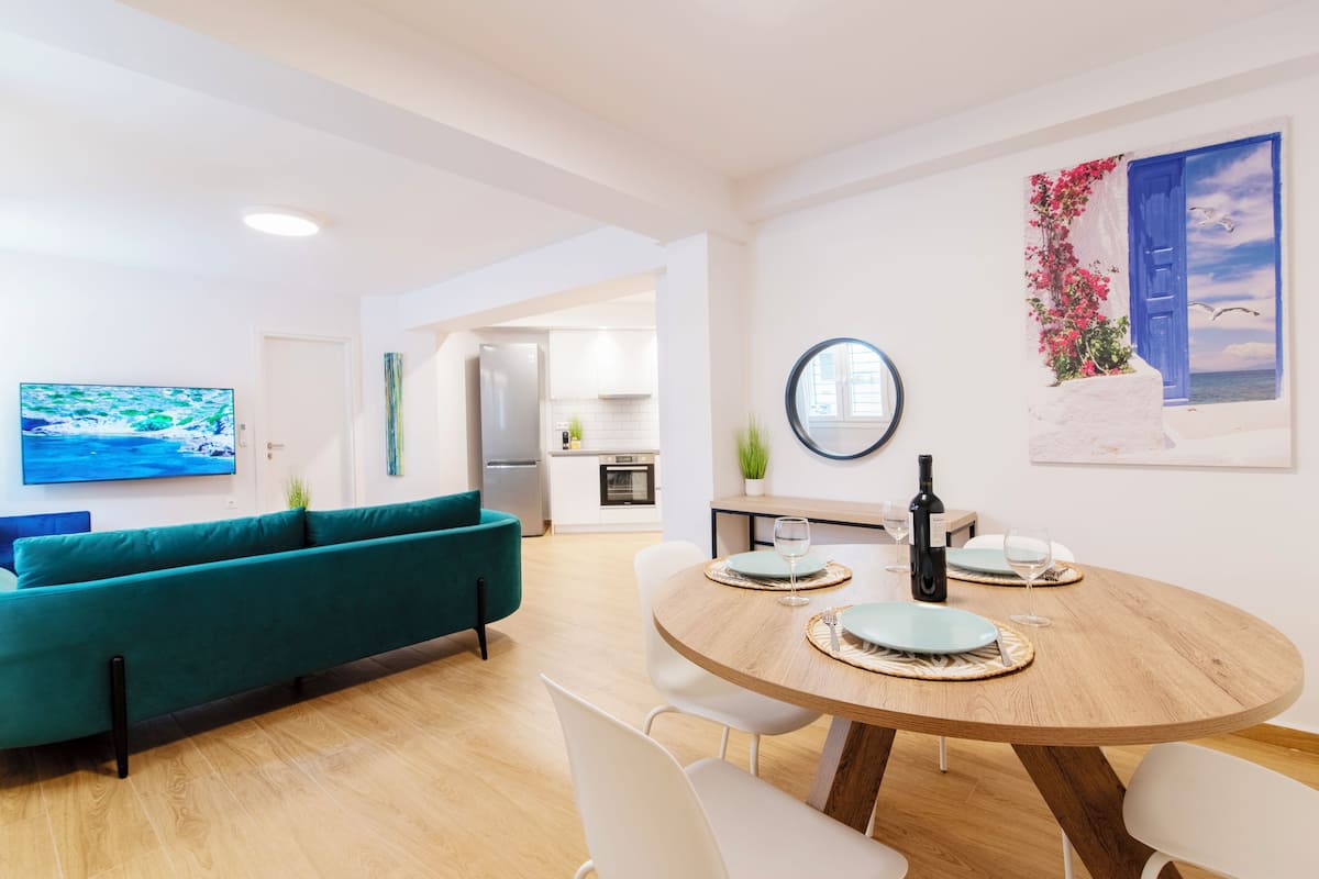 A modern living room with a teal sofa, a wooden dining table with four chairs, and a kitchen in the background.