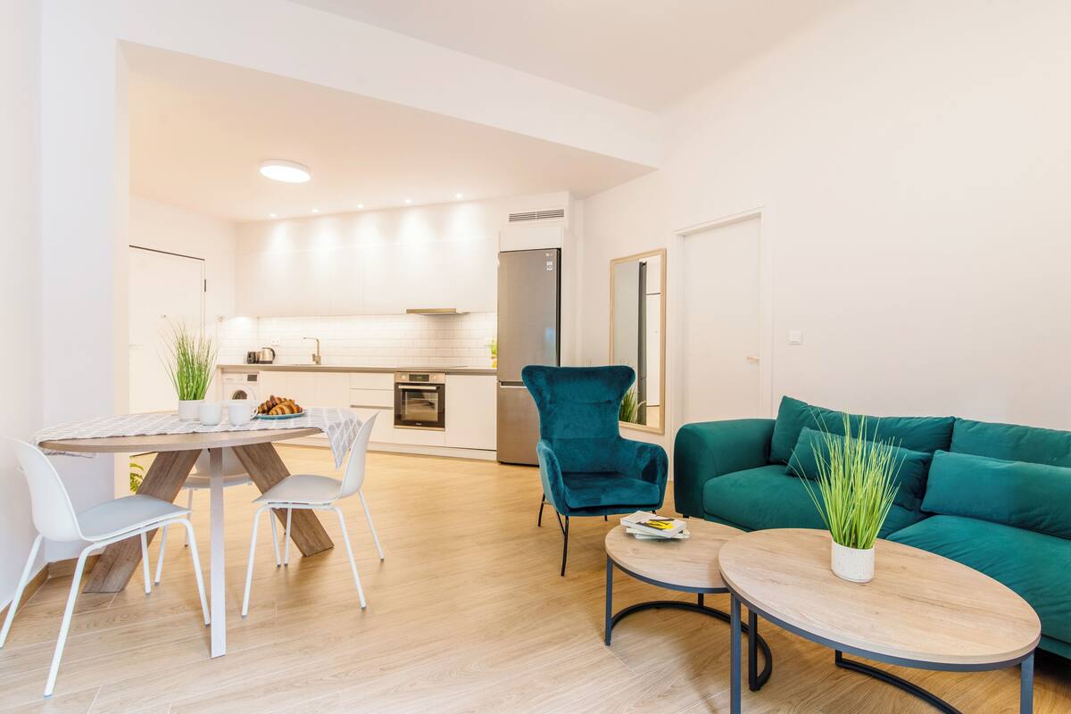A modern living room with a green sofa, a dining table with chairs, and a kitchen area in the background.