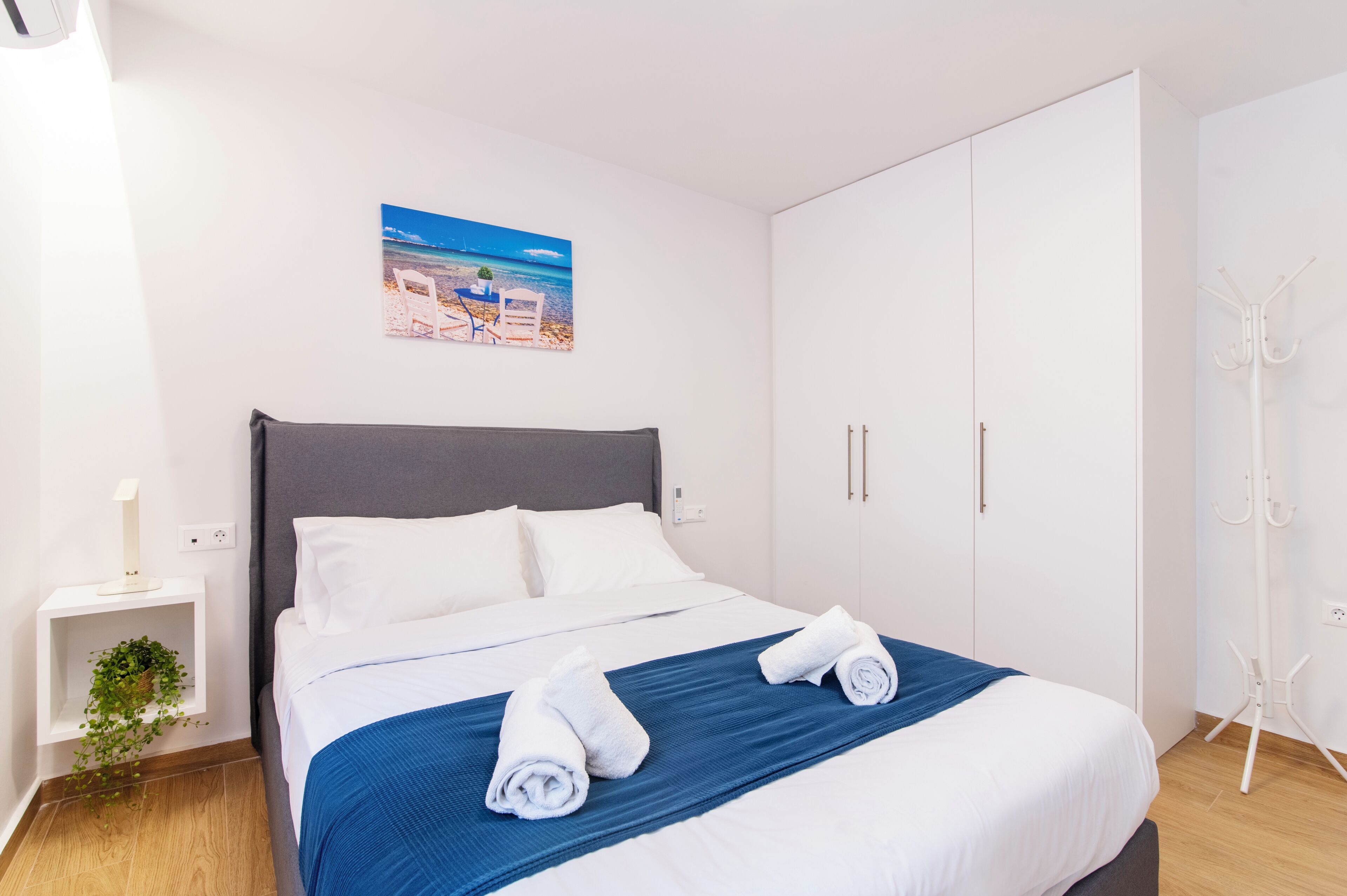 Een opgemaakt bed met witte en blauwe beddengoed, een nachtkastje met een lamp, en een wandgemonteerde kapstok.