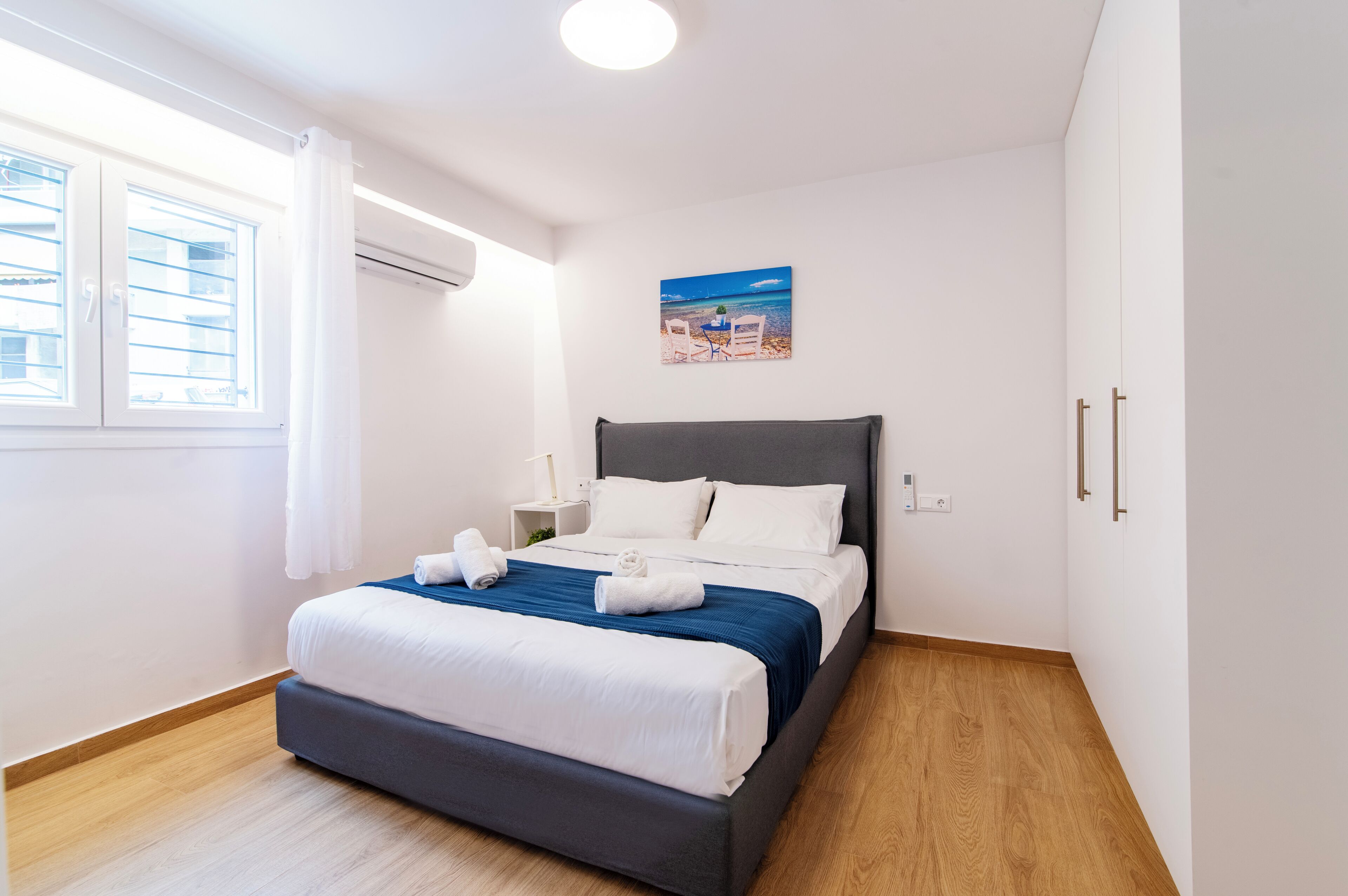 A neatly made bed with white and blue bedding, a nightstand with a lamp, and a beach scene painting on the wall.