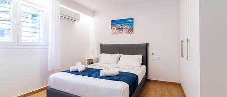 A neatly made bed with white and blue bedding, a nightstand with a lamp, and a beach scene painting on the wall.