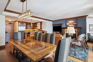 A spacious living area with a large wooden dining table, comfortable seating, a fireplace, and a kitchen in the background.