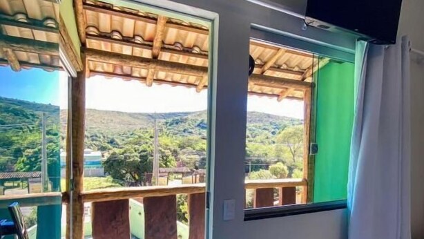 A room with a balcony, a TV mounted on the wall, and a view of a green landscape. - Pousada Cardeal Mota (Santana do Riacho)