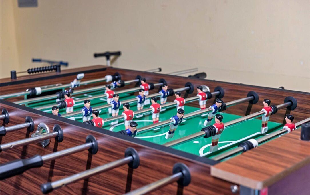 A foosball table with two teams, one in red and one in blue, in a room with a wooden floor and a beige wall.