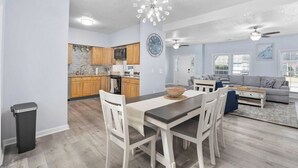A modern kitchen with wooden cabinets, a dining area with a table and chairs, and a living room with a sofa and a large window.