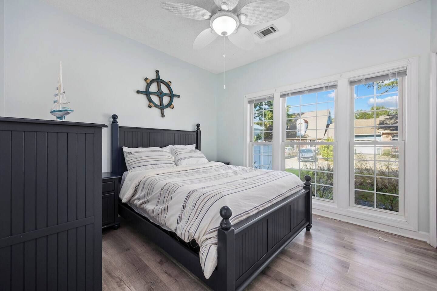 Une chambre à coucher comprenant un lit, une table de chevet, un ventilateur de plafond, un élément de décoration en forme de roue de navire, et une vue sur l’extérieur grâce à de grandes fenêtres.