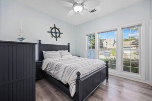 A bedroom with a bed, a nightstand, a ceiling fan, a ship's wheel decoration, and a view of the outside through large windows.