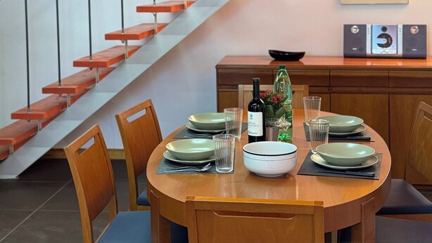 Un comedor con una mesa de madera dispuesta para cuatro, una escalera con escalones de madera y una planta colgante.