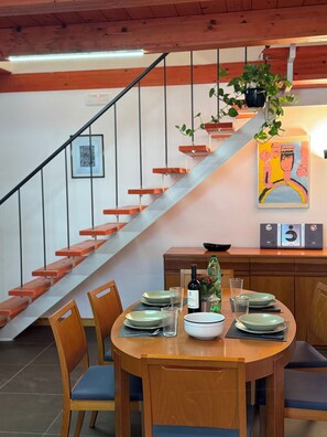 A dining area with a wooden table set for four, a staircase with wooden steps, and a hanging plant. - Casa Claudia (Salerno)