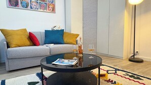 A living room with a grey sofa, colorful cushions, a round coffee table with magazines, a glass of wine, and a patterned rug.